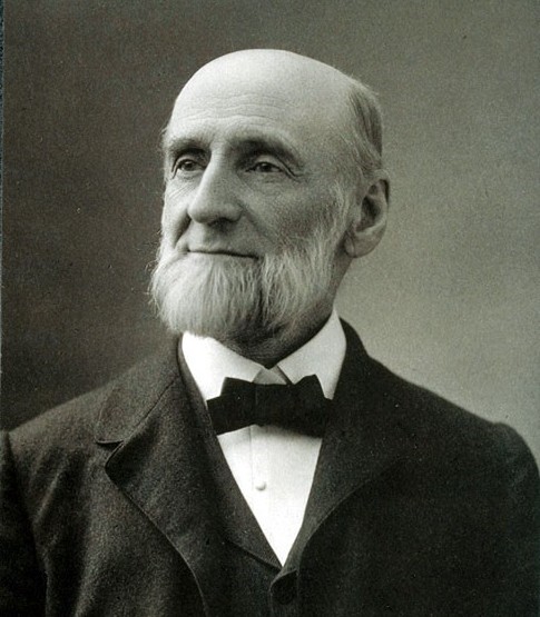 Black and white studio portrait of Gamaliel Bradford, a balding, bearded old man facing the camera but looking sideways in the direction the light is coming from. He looks healthy and hardy. He is wearing a dark suit with vest, white shirt, and black bowtie. The style of clothing appears to be circa 1900. He appears to be about 75 years old, looks contented, and is smiling.