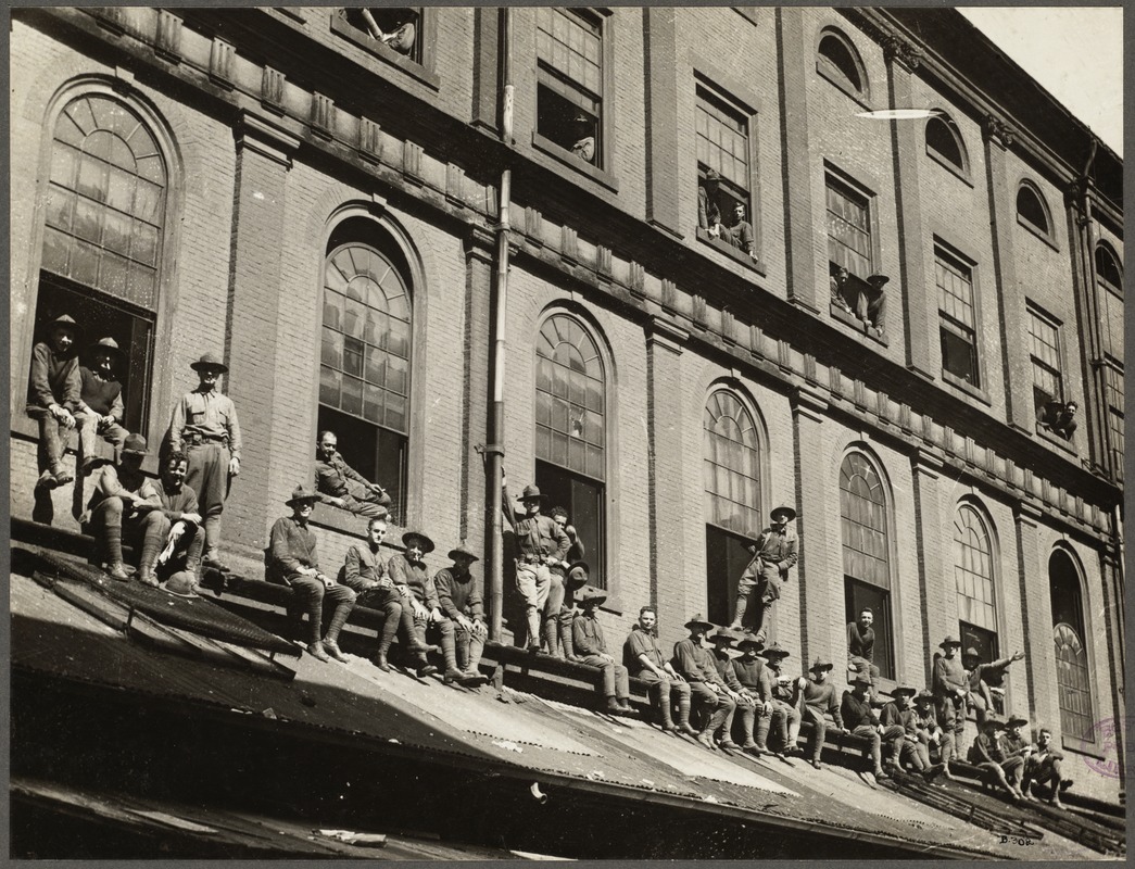 Black and white photograph of a group of State Guards posing for the camera. The photograph features the outside of the upper two levels of Faneuil Hall. There is a line of guards sitting on top of a roof that is coming out of the side of the Hall just over the first floor. Behind this line of men are a few standing, and a few sitting in or leaning out of the second floor windows. There is also a group of guards leaning out of the third floor windows. The men are wearing State Guard uniforms, including khaki shirts, khaki breeches or pants, long socks, shoes, and campaign hats. The nature of the photograph is relatively jovial, some of the men are smiling, one man is lying down on his side in a second story window, another is standing on a window sill and leaning out of the window, and a third has his arm out as if waving to the photographer.