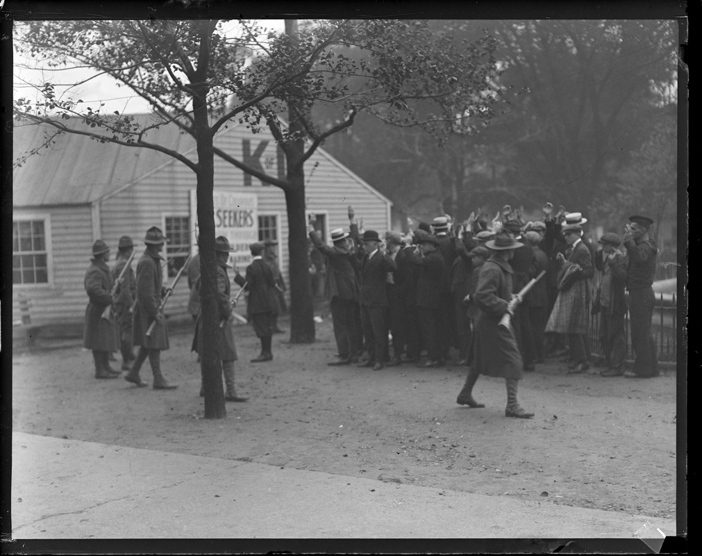 Black and white photograph of people gathered in front of a small, one story building. The building has light colored vinyl siding and has two large letters on the front of it above two windows and a door. One of the letters is K, and the other is obscured by a tree. There is also a sign on the front of the building. The only legible word on the sign says "seekers." Most of the people featured in the photograph are men and boys gathered between the building on the left and a short metal fence on the right half of the image. They are all wearing dark suits and have hats, some of which are boaters, newsboys, and bowlers. One person at the front seems to be a younger man wearing breeches. These people are gathered tightly together facing towards the building and all have their hands up. They are surrounded by a group of a smaller number of State Guard officials, the majority of which are on the left side of the image. They are in State Guard uniforms including trench coats and campaign hats. They each hold a rifle at the ready by their side. They are pointing the rifles towards the larger group of men and boys that they are surrounding.